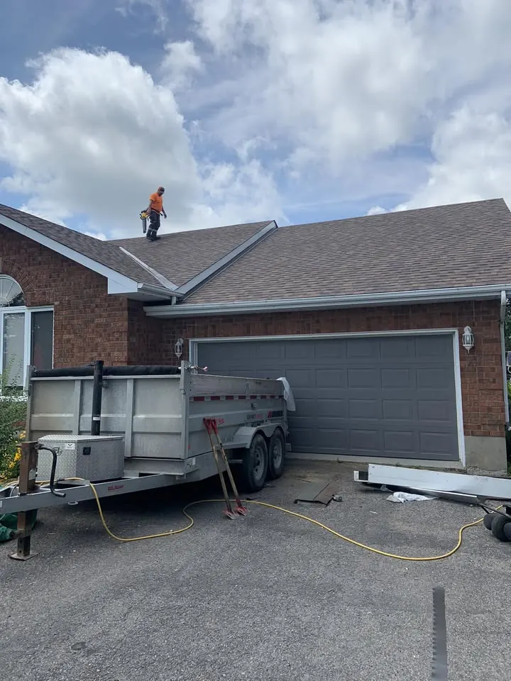 Brick house with a new grey shingle roof and white eavestrough, a dump trailer in the driveway and a roofer on the slope