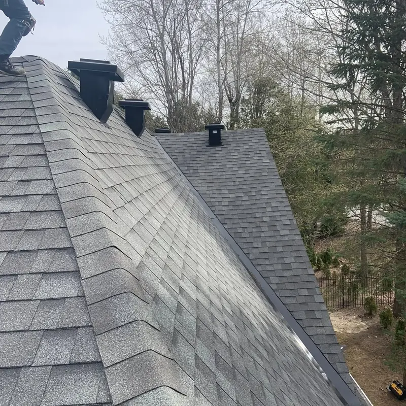 Newly shingled roof slope with two black vent stacks and a ridge, bare trees behind